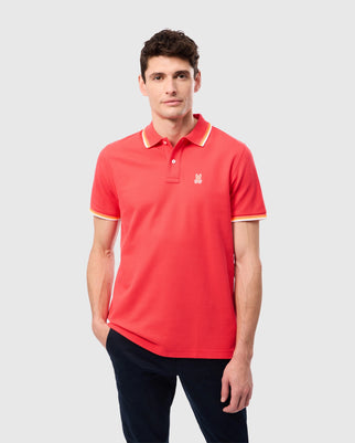 A man confidently stands against a plain backdrop, wearing the Psycho Bunny MENS GRANBURY PIQUE POLO - B6K400C200 in bright pink with yellow-trimmed sleeves and collar, paired with dark pants. He sports short dark hair and casually rests his left hand in his pocket while gazing forward.