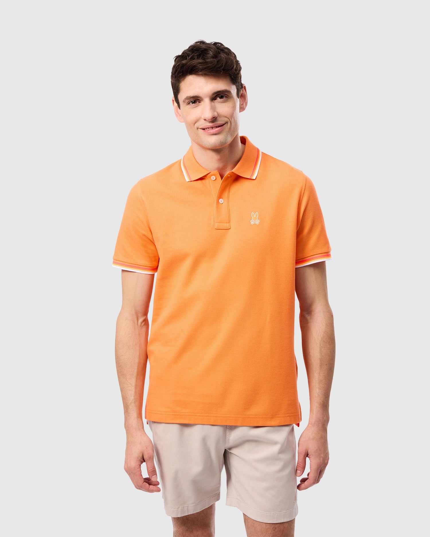 A young man smiles at the camera, wearing the Psycho Bunny MENS GRANBURY PIQUE POLO - B6K400C200 in orange, featuring a rib knit collar and white trim, paired with beige shorts against a plain light background.