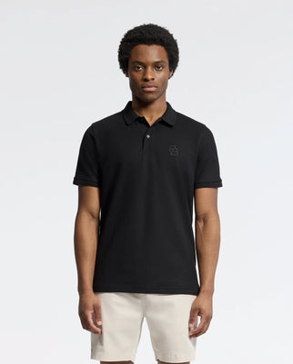A person is wearing Psycho Bunny's classic black Nicholson Rib Collar Pique Polo (B6K401F200), made of soft Pima cotton with an embroidered logo, paired with beige shorts. They stand against a plain backdrop, hair styled naturally, looking directly at the camera.