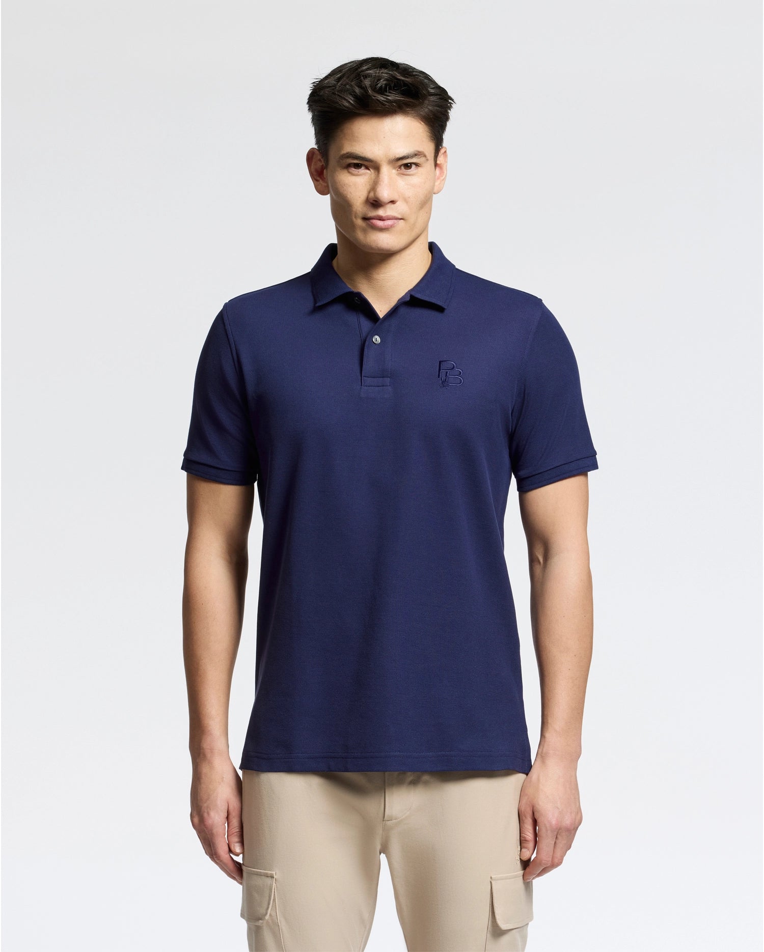 A man stands against a plain background wearing the Psycho Bunny MENS NICHOLSON RIB COLLAR PIQUE POLO in navy, crafted from luxurious Pima cotton, and beige pants. The shirt features an embroidered logo on the chest. He has short dark hair and a neutral expression.