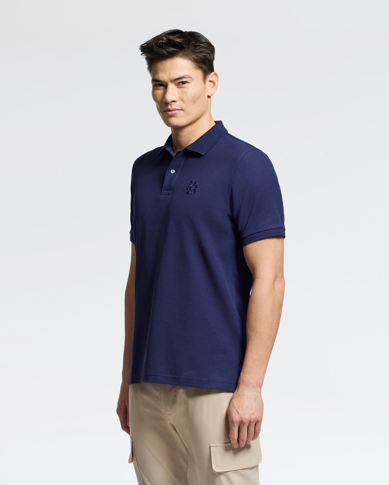 A person with short dark hair is wearing a navy blue Psycho Bunny men's Nicholson rib collar pique polo, subtly adorned with an embroidered logo, paired with beige cargo pants. They stand against a light background, looking slightly to the side.