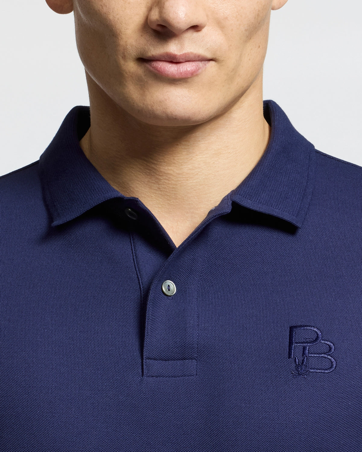 A person wearing Psycho Bunny's dark blue polo, the Nicholson Rib Collar Pique Polo - B6K401F200, made from premium Pima cotton with a small embroidered logo on the chest. The focus is on the upper torso, emphasizing the collar and buttons against a light background.