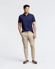 A person stands with hands in pockets, wearing a Psycho Bunny navy blue Nicholson Rib Collar Pique Polo (B6K401F200), beige cargo pants, and sneakers against a plain light-colored background.