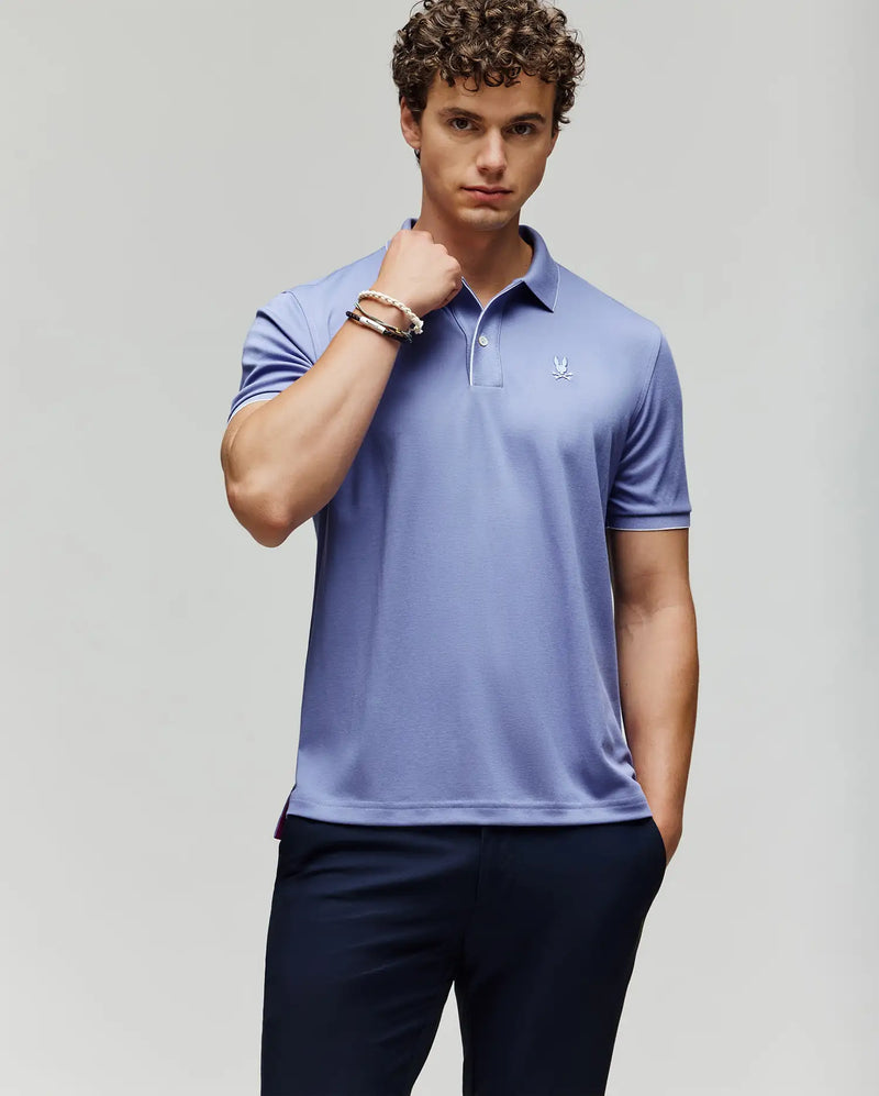 A young man with curly hair wears the Psycho Bunny MENS BRYCE PERFORMANCE PIQUE POLO - B6K486F200 in light purple and dark pants, standing against a plain background with one hand in his pocket and the other touching his collar.