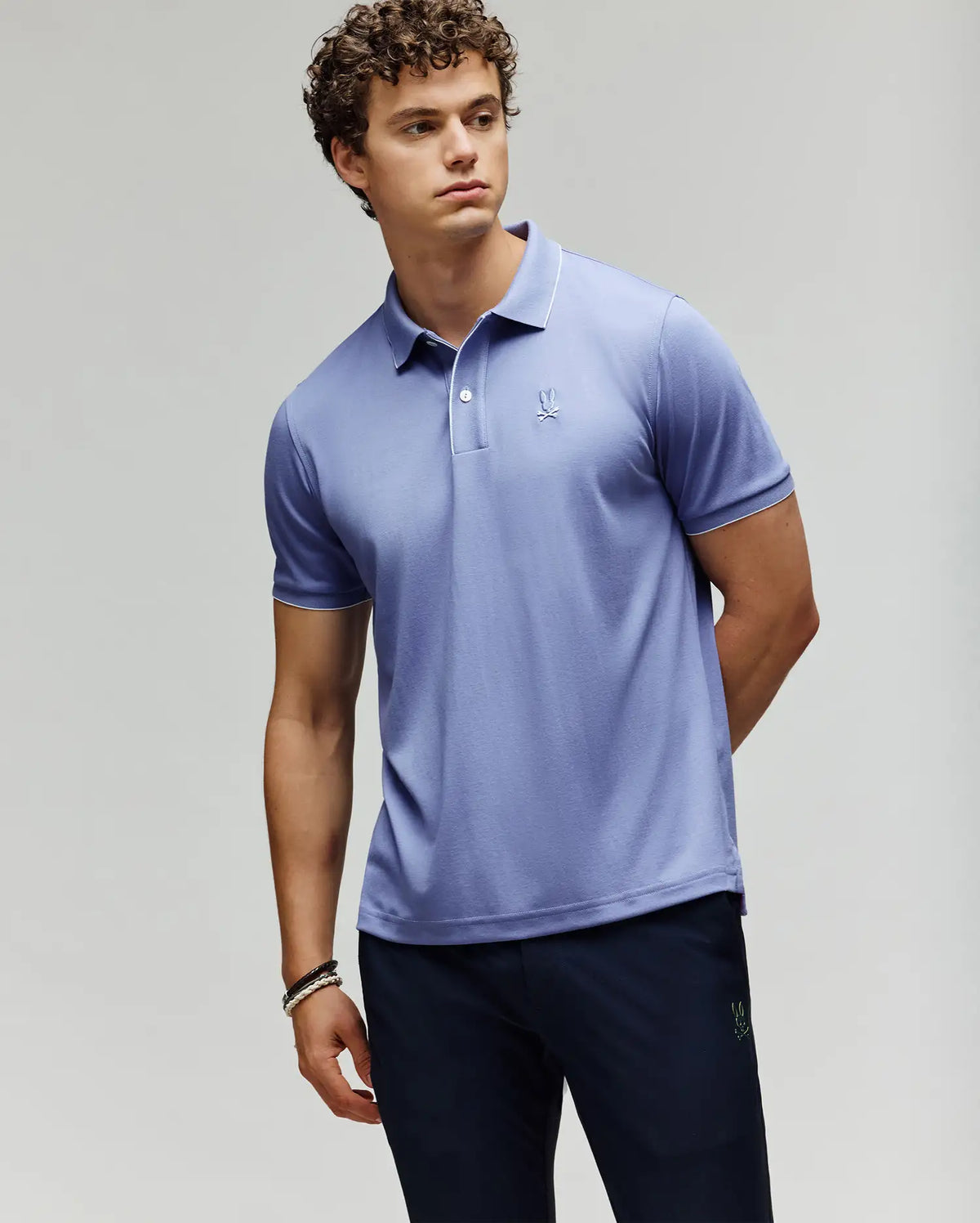 A young man wearing a Psycho Bunny MENS BRYCE PERFORMANCE PIQUE POLO (B6K486F200) in light purple and dark pants stands against a plain background, arms relaxed—one behind his back—looking off to the side.
