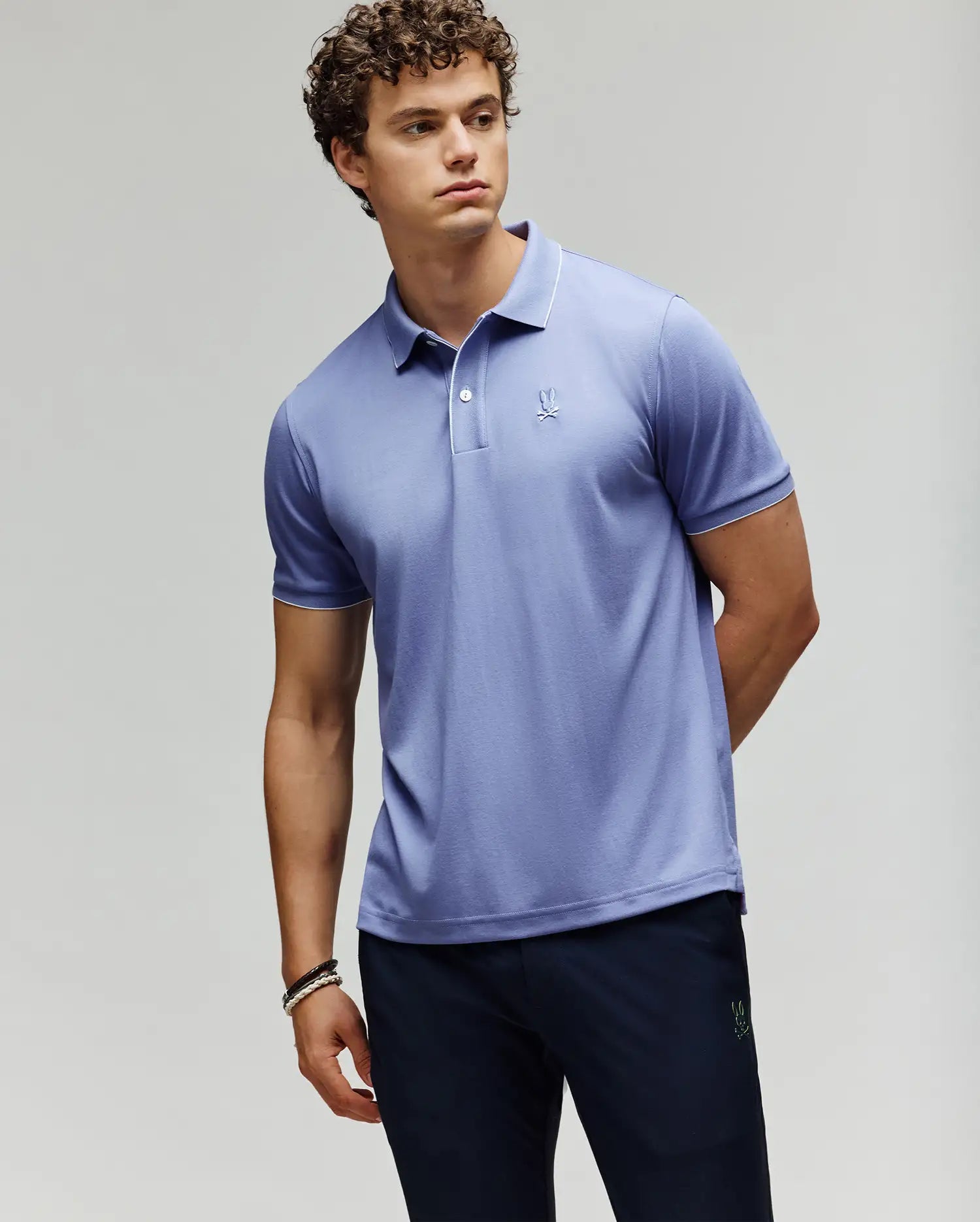 A young man wearing a Psycho Bunny MENS BRYCE PERFORMANCE PIQUE POLO (B6K486F200) in light purple and dark pants stands against a plain background, arms relaxed—one behind his back—looking off to the side.