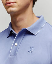 A close-up of a person in the Psycho Bunny MENS BRYCE PERFORMANCE PIQUE POLO (B6K486F200), light blue pima cotton with white piping on the collar and an embroidered bunny head and crossbones logo. The face is partially out of frame.
