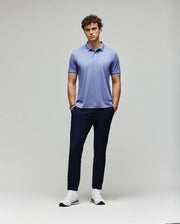 A man with curly hair stands against a plain background, wearing Psycho Bunny's MENS BRYCE PERFORMANCE PIQUE POLO (B6K486F200) in light purple, with dark navy pants and white sneakers. His hands are in his pockets and he has a neutral expression.