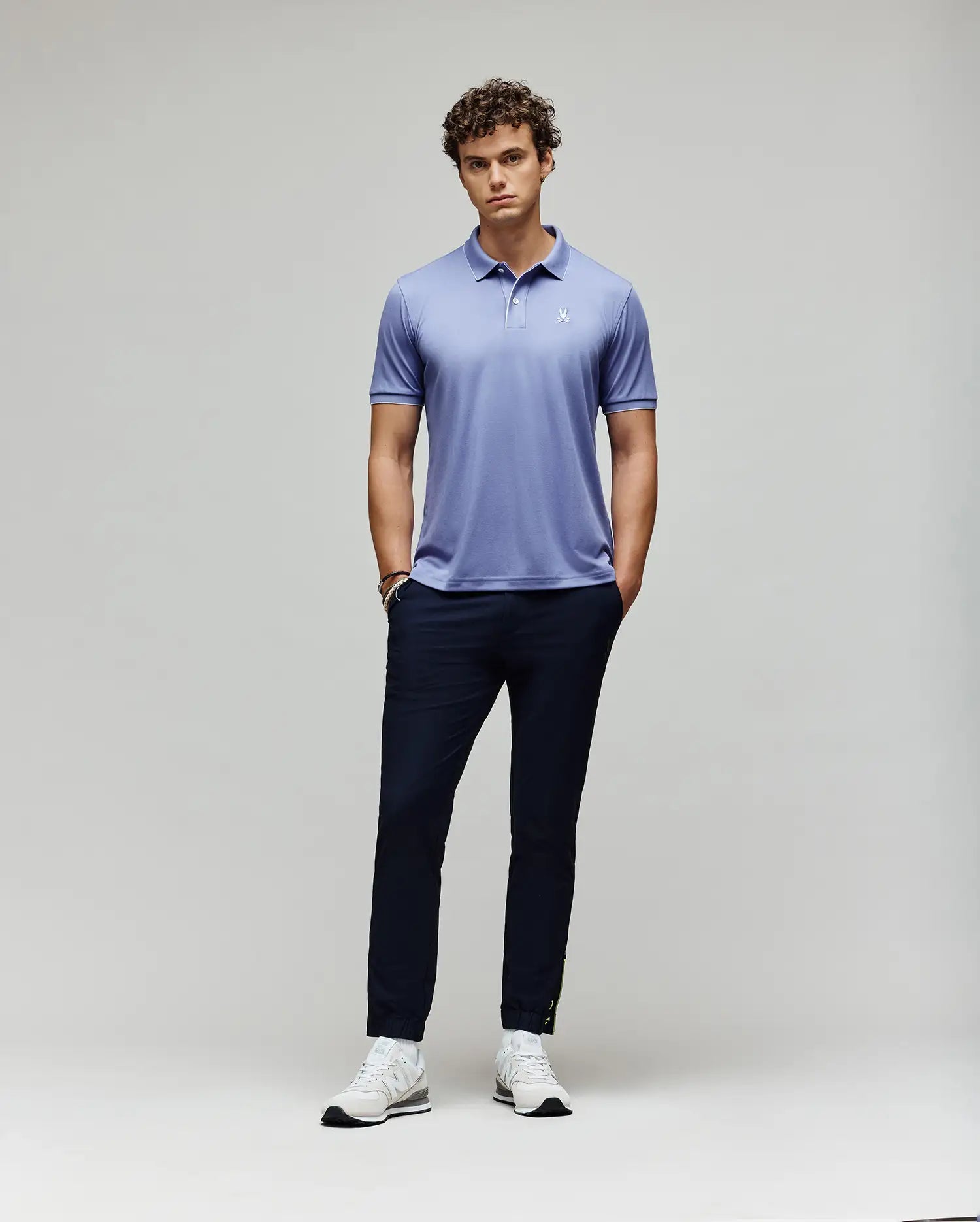 A man with curly hair stands against a plain background, wearing Psycho Bunny's MENS BRYCE PERFORMANCE PIQUE POLO (B6K486F200) in light purple, with dark navy pants and white sneakers. His hands are in his pockets and he has a neutral expression.