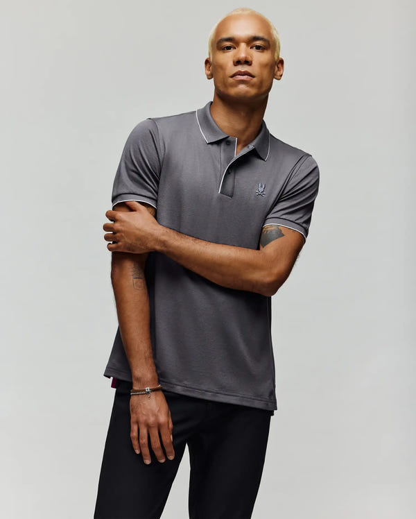 A man with short blond hair wears a Psycho Bunny MENS BRYCE PERFORMANCE PIQUE POLO SHIRT (B6K486F200) in gray and black pants, standing against a plain light background with one arm crossed and a neutral expression.