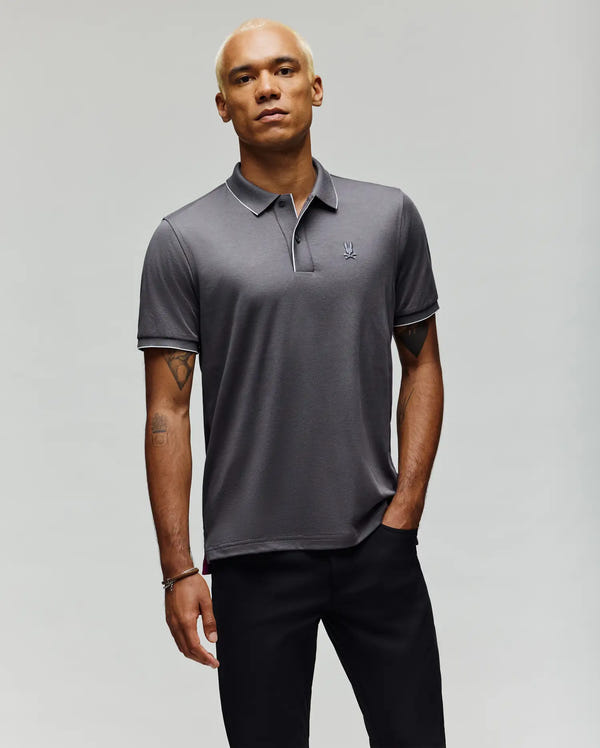 A man with short, bleached blond hair wears the Psycho Bunny MENS BRYCE PERFORMANCE PIQUE POLO SHIRT (B6K486F200) in grey, featuring a small chest logo, paired with black pants, standing with one hand in his pocket against a plain light background.