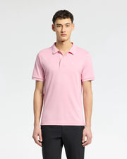 A person in a Psycho Bunny MENS BRYCE PERFORMANCE PIQUE POLO, crafted from breathable moisture-wicking fabric, stands against a white background. With short dark hair and black pants, their relaxed arms highlight the polo's comfort and style.
