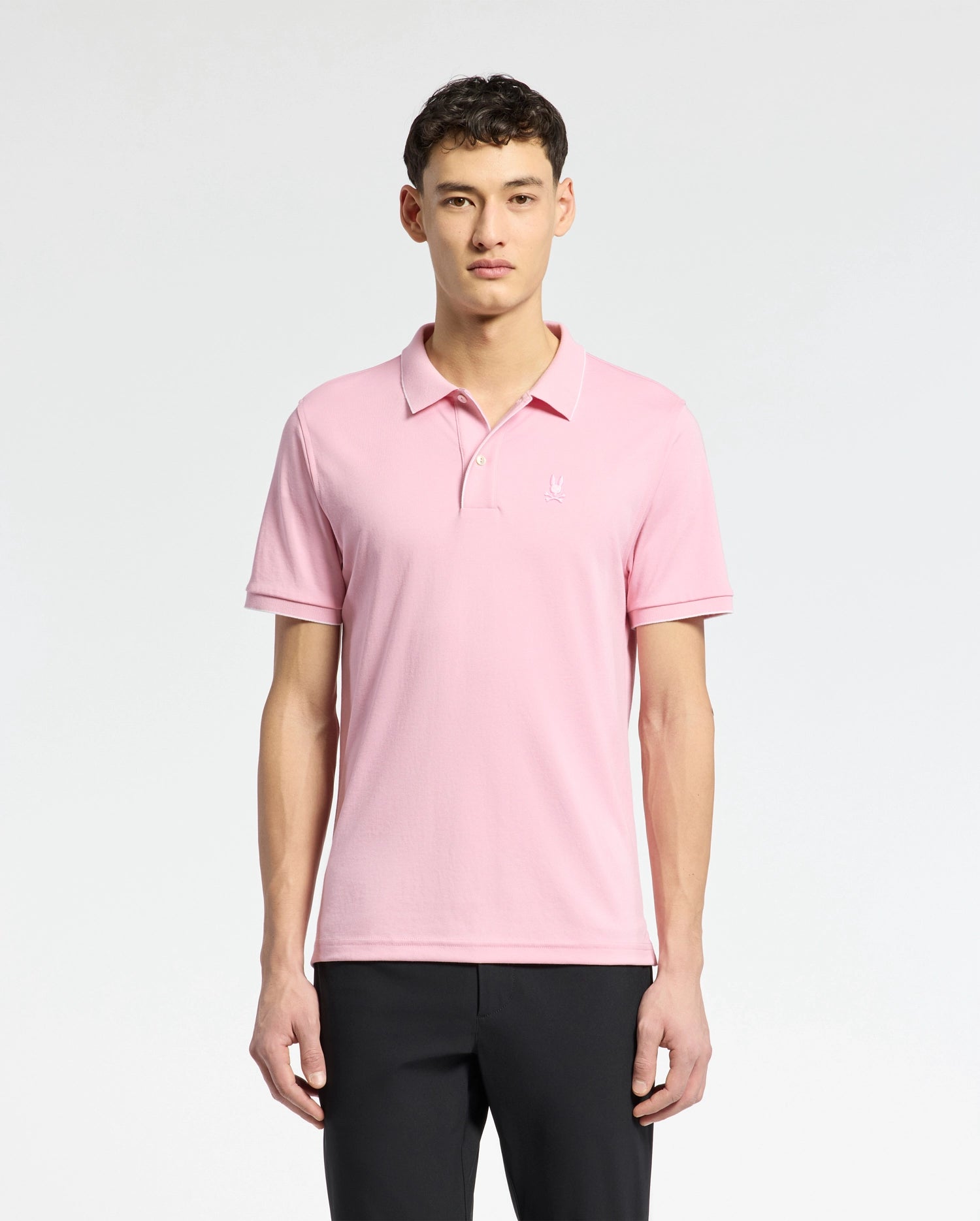 A person in a Psycho Bunny MENS BRYCE PERFORMANCE PIQUE POLO, crafted from breathable moisture-wicking fabric, stands against a white background. With short dark hair and black pants, their relaxed arms highlight the polo's comfort and style.