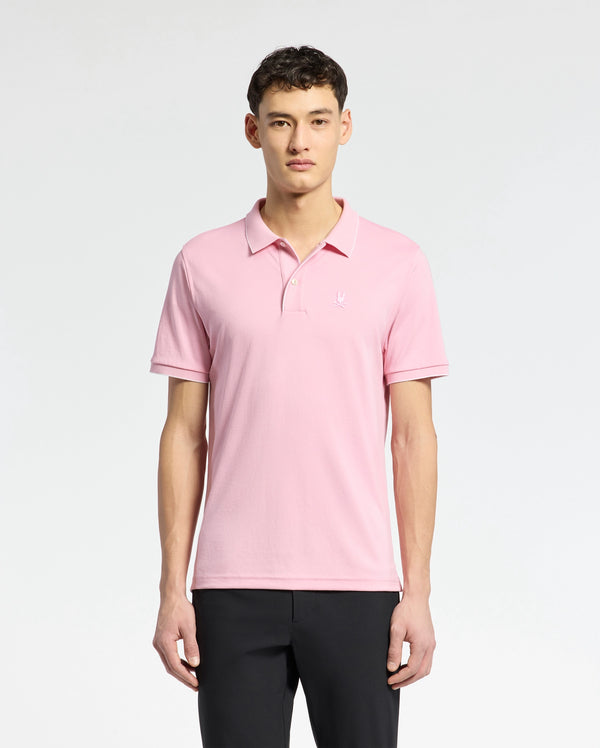 A person in a Psycho Bunny MENS BRYCE PERFORMANCE PIQUE POLO, crafted from breathable moisture-wicking fabric, stands against a white background. With short dark hair and black pants, their relaxed arms highlight the polo's comfort and style.
