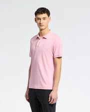 Against a gray background, a man wears the Psycho Bunny MENS BRYCE PERFORMANCE PIQUE POLO - B6K486F200 in light pink, made from breathable moisture-wicking fabric. The shirt has an embroidered logo on the chest and pairs well with his black pants. His short curly hair completes the elegant look.