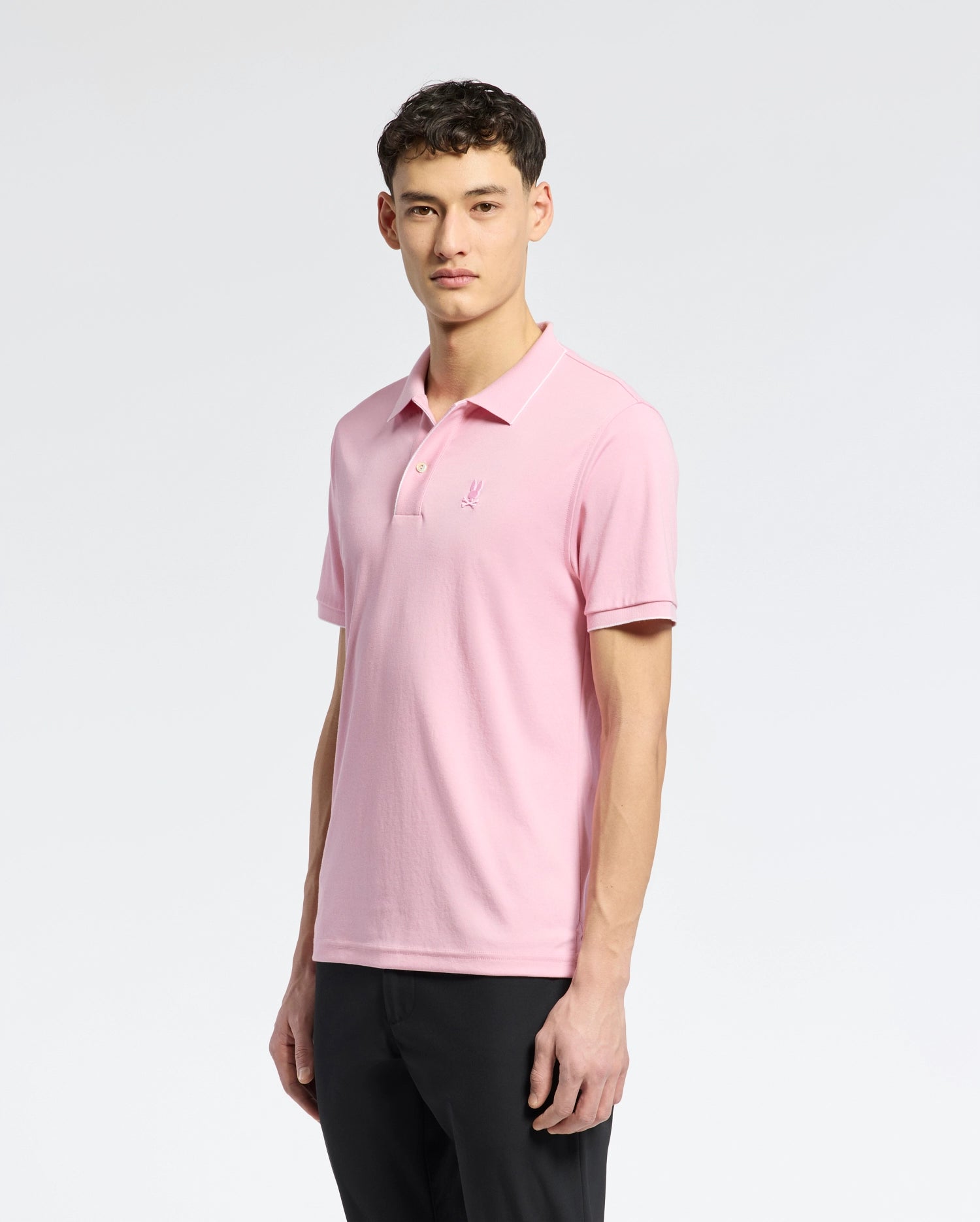 Against a gray background, a man wears the Psycho Bunny MENS BRYCE PERFORMANCE PIQUE POLO - B6K486F200 in light pink, made from breathable moisture-wicking fabric. The shirt has an embroidered logo on the chest and pairs well with his black pants. His short curly hair completes the elegant look.