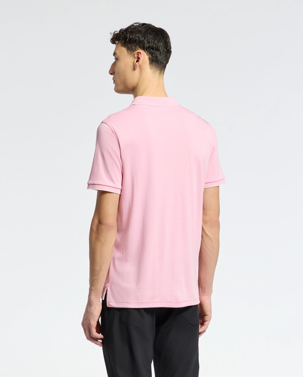 A man with short, dark hair faces away, wearing Psycho Bunny's Mens Bryce Performance Pique Polo - B6K486F200 in light pink made from breathable moisture-wicking fabric and black pants against a plain white background.