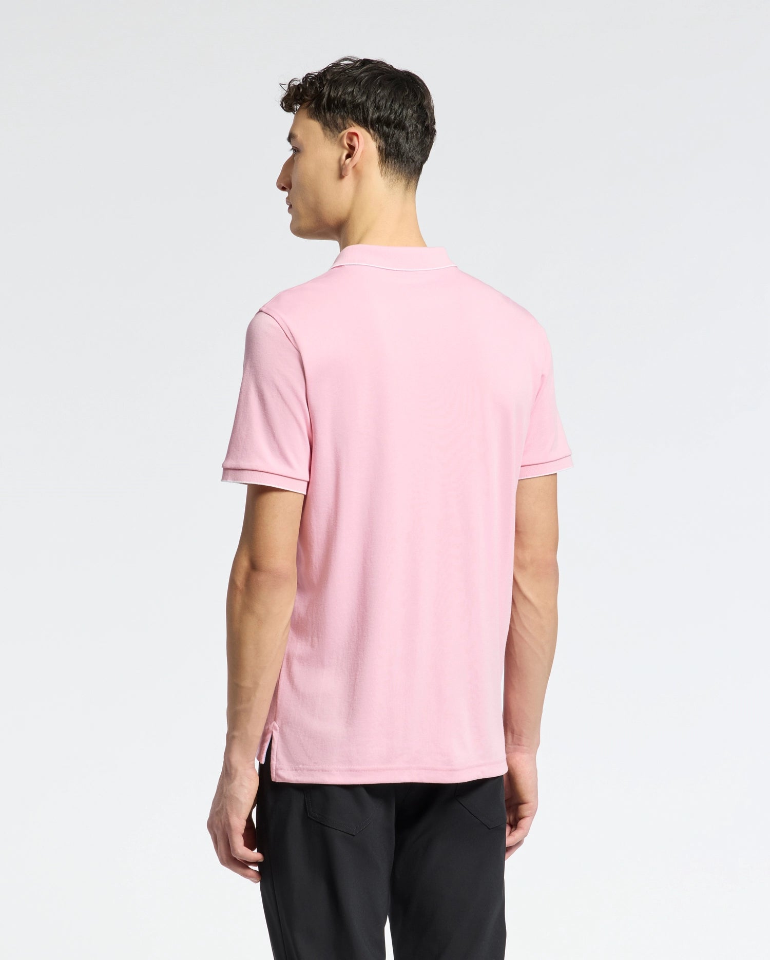 A man with short, dark hair faces away, wearing Psycho Bunny's Mens Bryce Performance Pique Polo - B6K486F200 in light pink made from breathable moisture-wicking fabric and black pants against a plain white background.