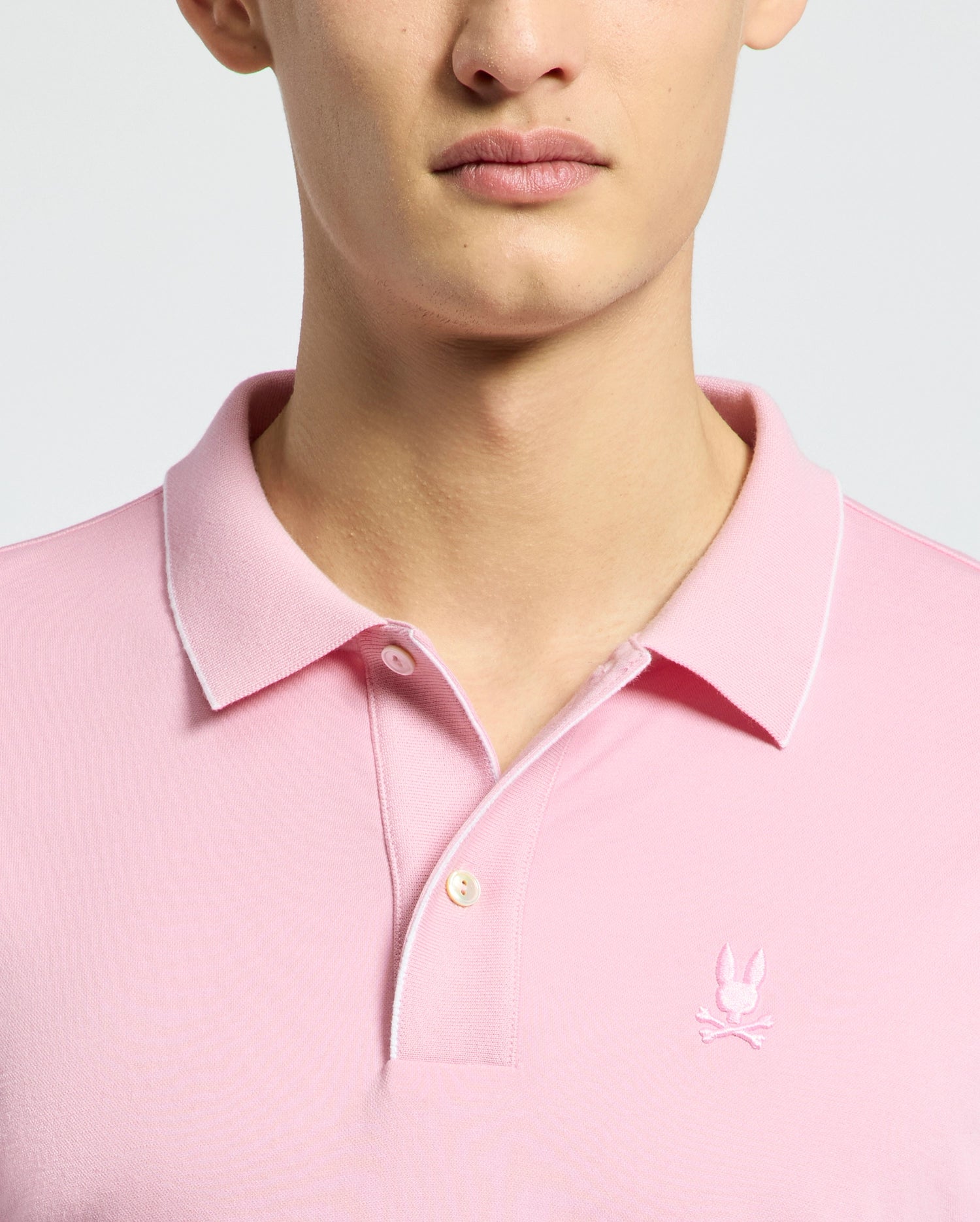 A person is wearing a Psycho Bunny MENS BRYCE PERFORMANCE PIQUE POLO (B6K486F200) in light pink, with an embroidered bunny and crossed bones on the left. It features a collar, two buttons, and breathable moisture-wicking fabric against a plain light gray background.