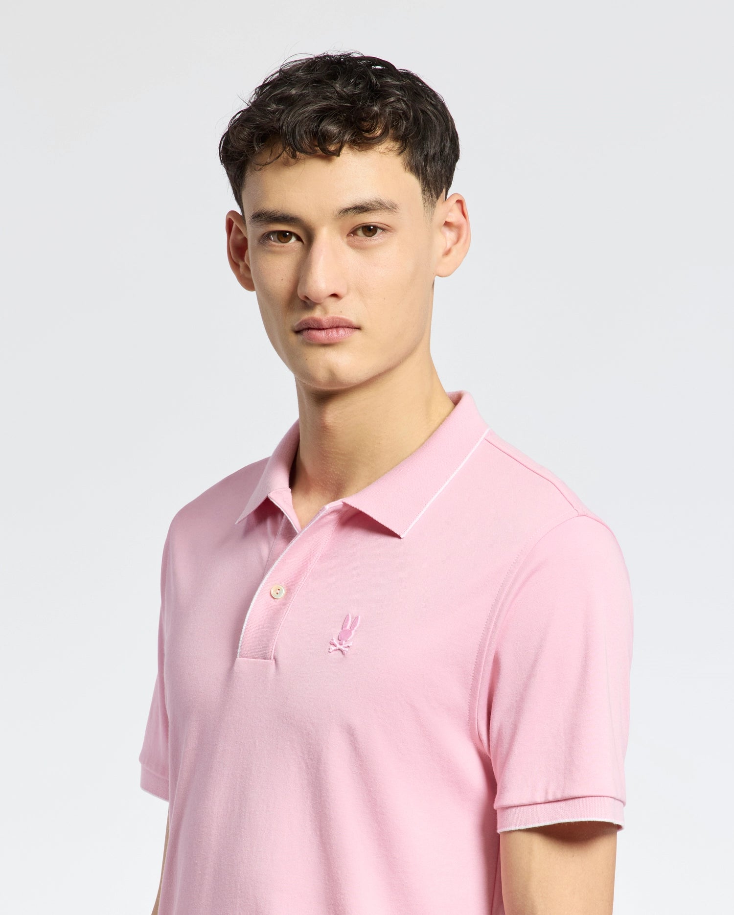 The individual, with short dark hair and a neutral expression, wears the Psycho Bunny MENS BRYCE PERFORMANCE PIQUE POLO in light pink, made from breathable moisture-wicking fabric, featuring a small embroidered logo on the left chest against a plain white backdrop.