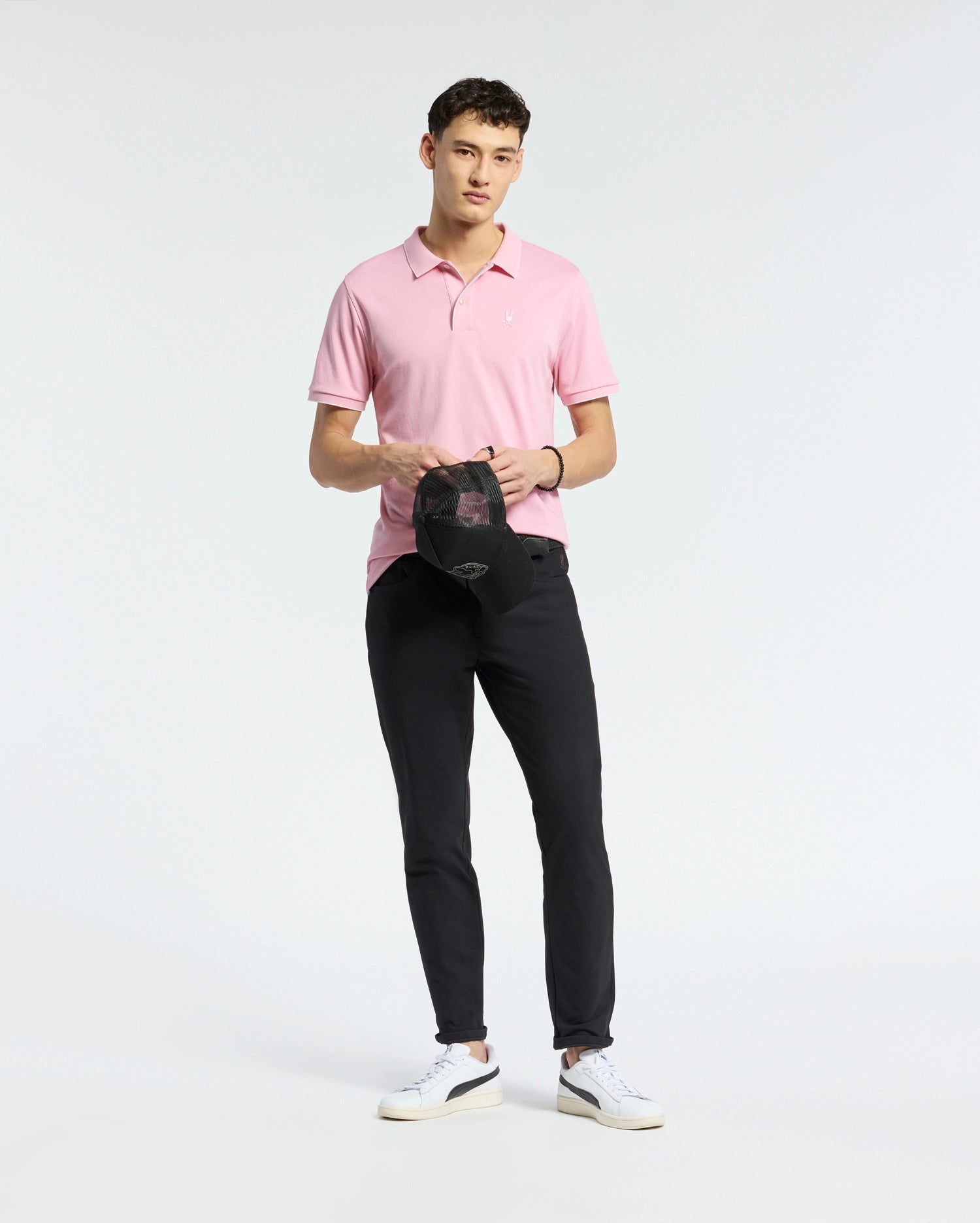 Wearing a Psycho Bunny light pink MENS BRYCE PERFORMANCE PIQUE POLO - B6K486F200, paired with black pants and white sneakers, a person holds a black cap against a plain white background.