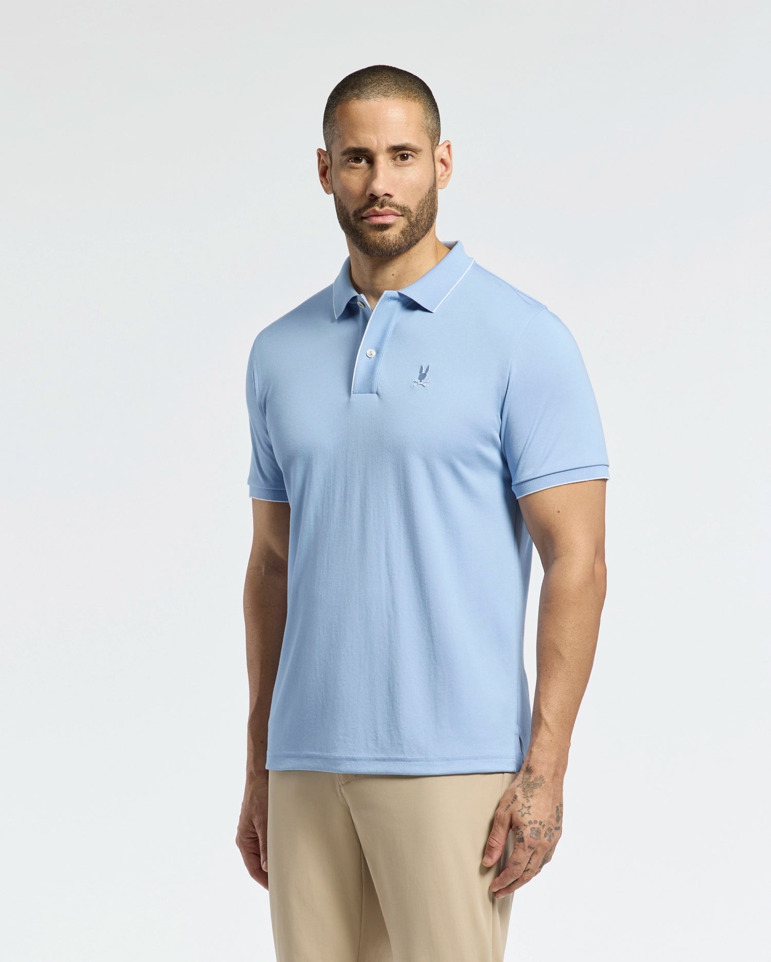 A bearded man wears the Psycho Bunny Men's Bryce Performance Pique Polo in light blue, featuring a small left-side logo, and beige pants as he stands against a white background, highlighting the breathable fabric.