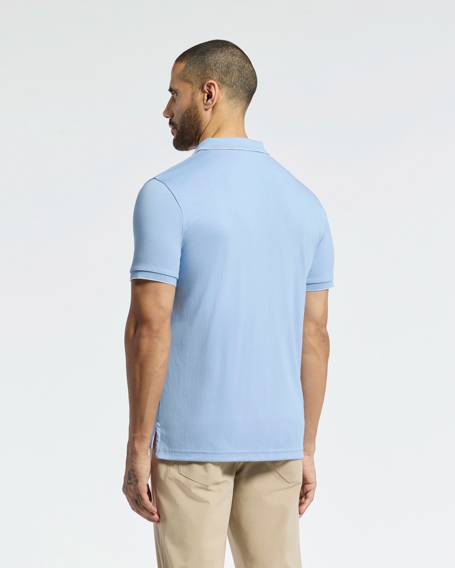 A man faces away from the camera in a light blue Psycho Bunny Bryce Performance Pique Polo (B6K486F200) and beige pants. This quick-dry shirt, made from breathable performance pique fabric, is set against a simple light grey background.