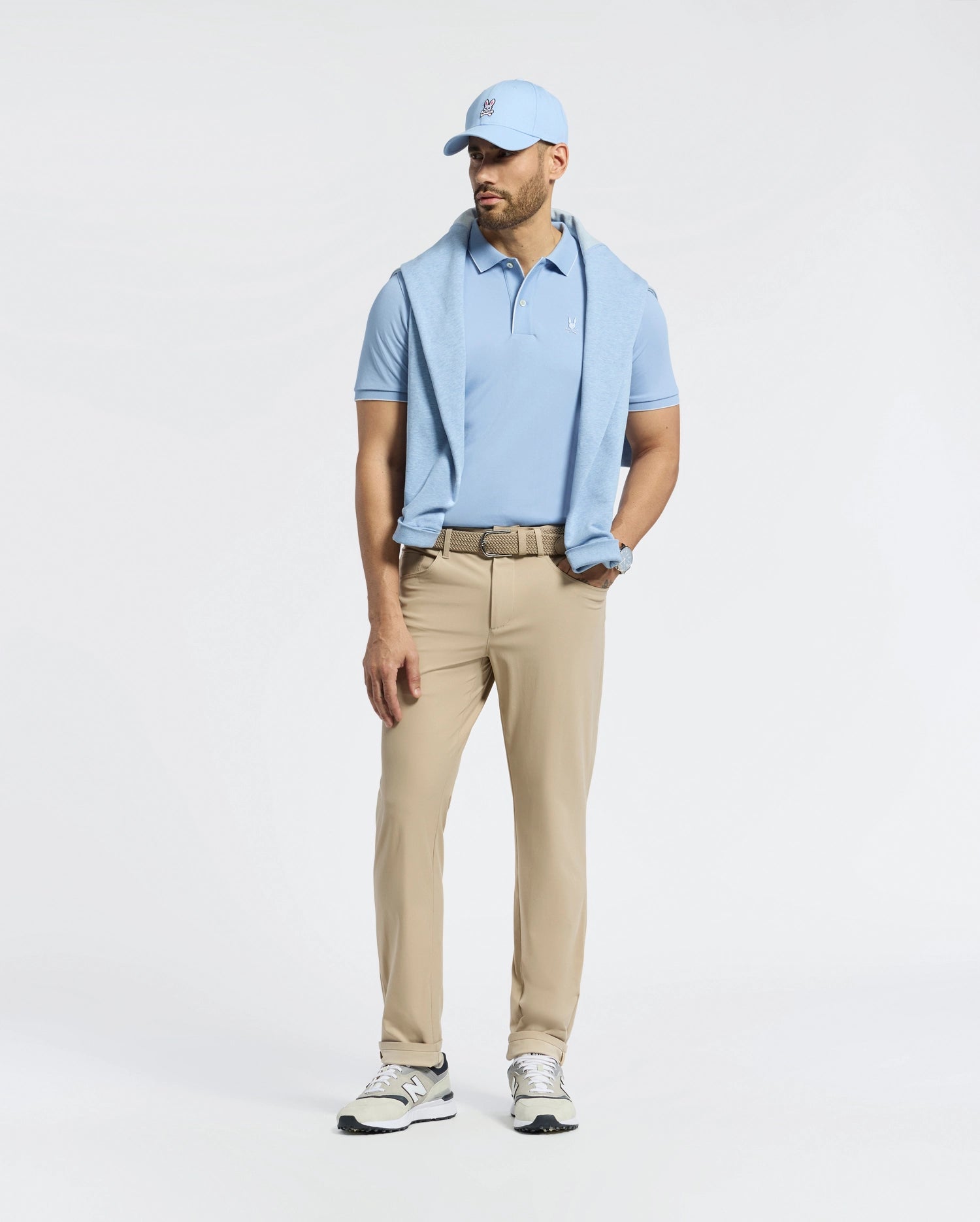 A man wearing a light blue Psycho Bunny Bryce Performance Pique Polo, matching baseball cap, and beige pants stands against a simple background. He has a light blue sweater draped over his shoulders and is wearing quick-dry white sneakers.