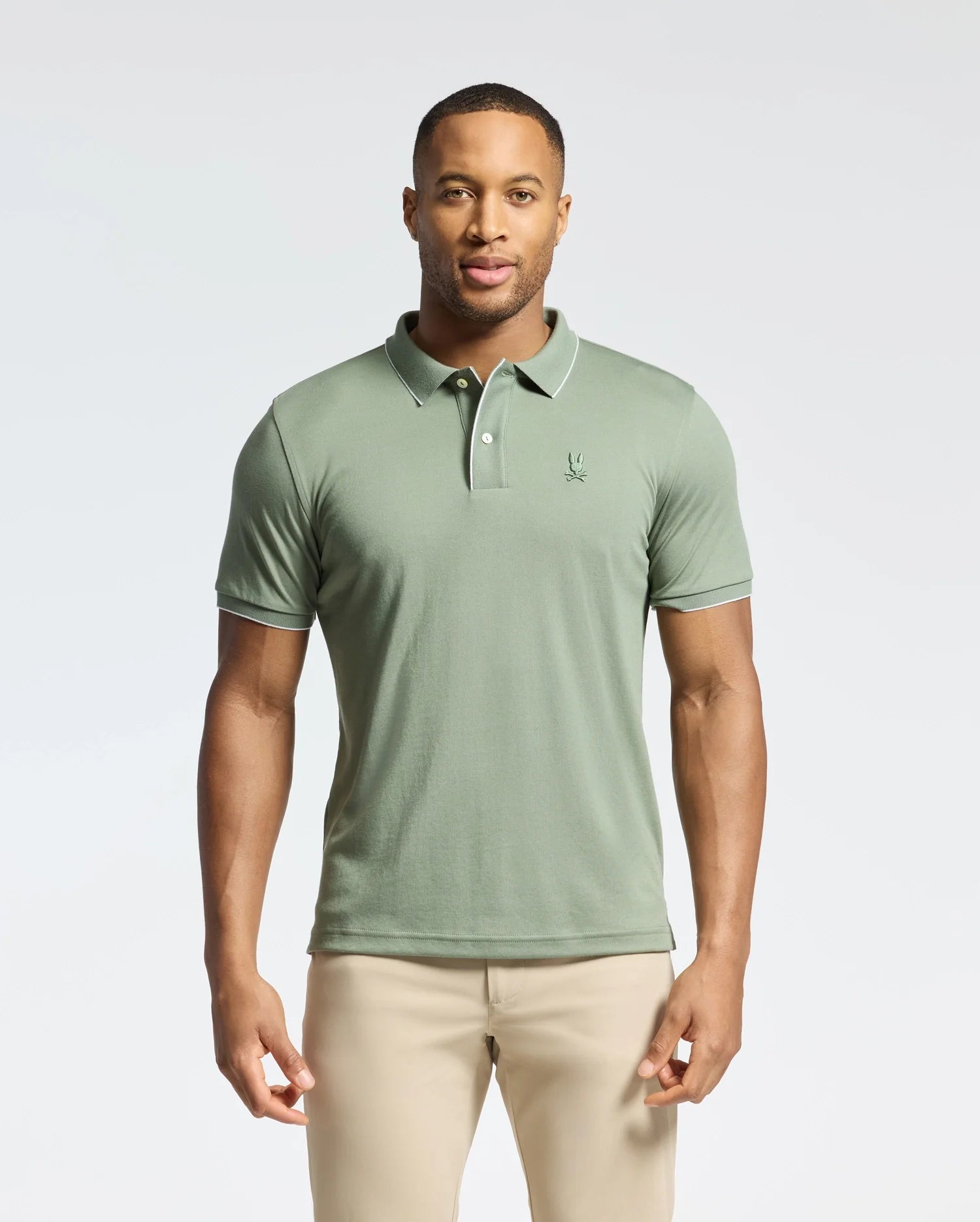 A man stands against a plain background wearing the Psycho Bunny MENS BRYCE PERFORMANCE PIQUE POLO (B6K486F200) in light green, made from breathable moisture-wicking fabric with a small embroidered logo on the chest, paired with beige pants.