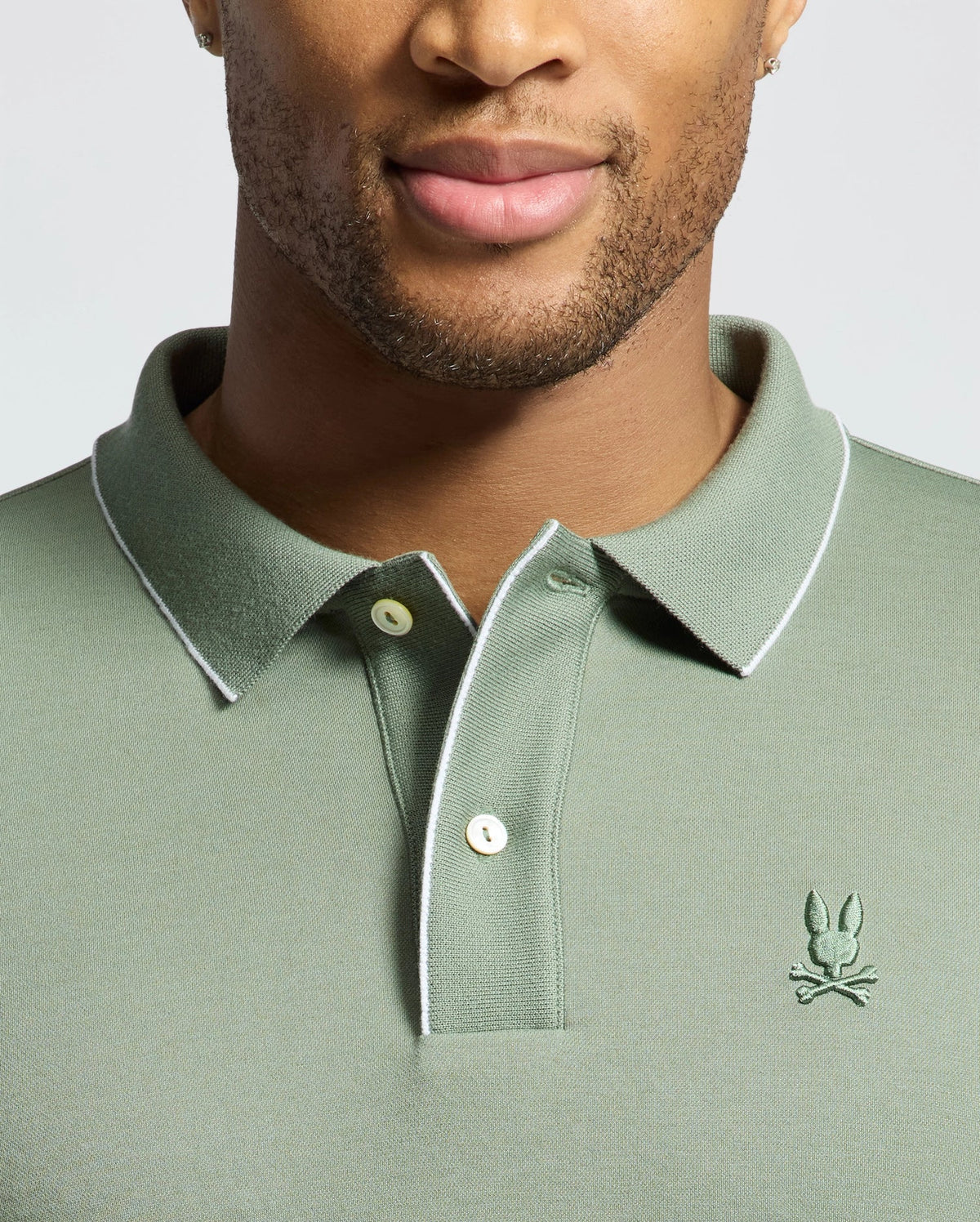 The focus is on a person wearing the Psycho Bunny MENS BRYCE PERFORMANCE PIQUE POLO (B6K486F200), featuring a green Modal-Pima cotton blend with white piping and embroidered chest logo, highlighting the breathable moisture-wicking fabric against a neutral background.