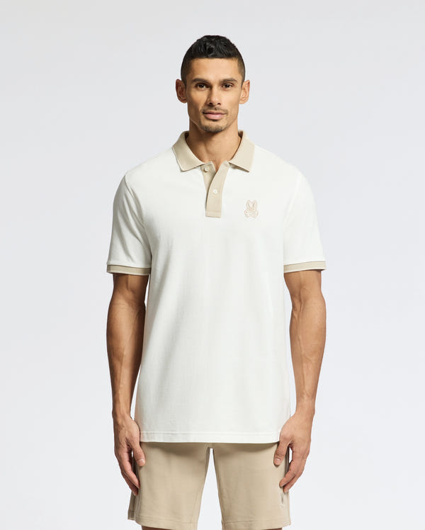 A man faces forward wearing the Psycho Bunny MENS NEW CASTLE PIQUE POLO - B6K487F200, a slim fit white polo with contrast beige collar detailing, paired with beige shorts against a plain light background.