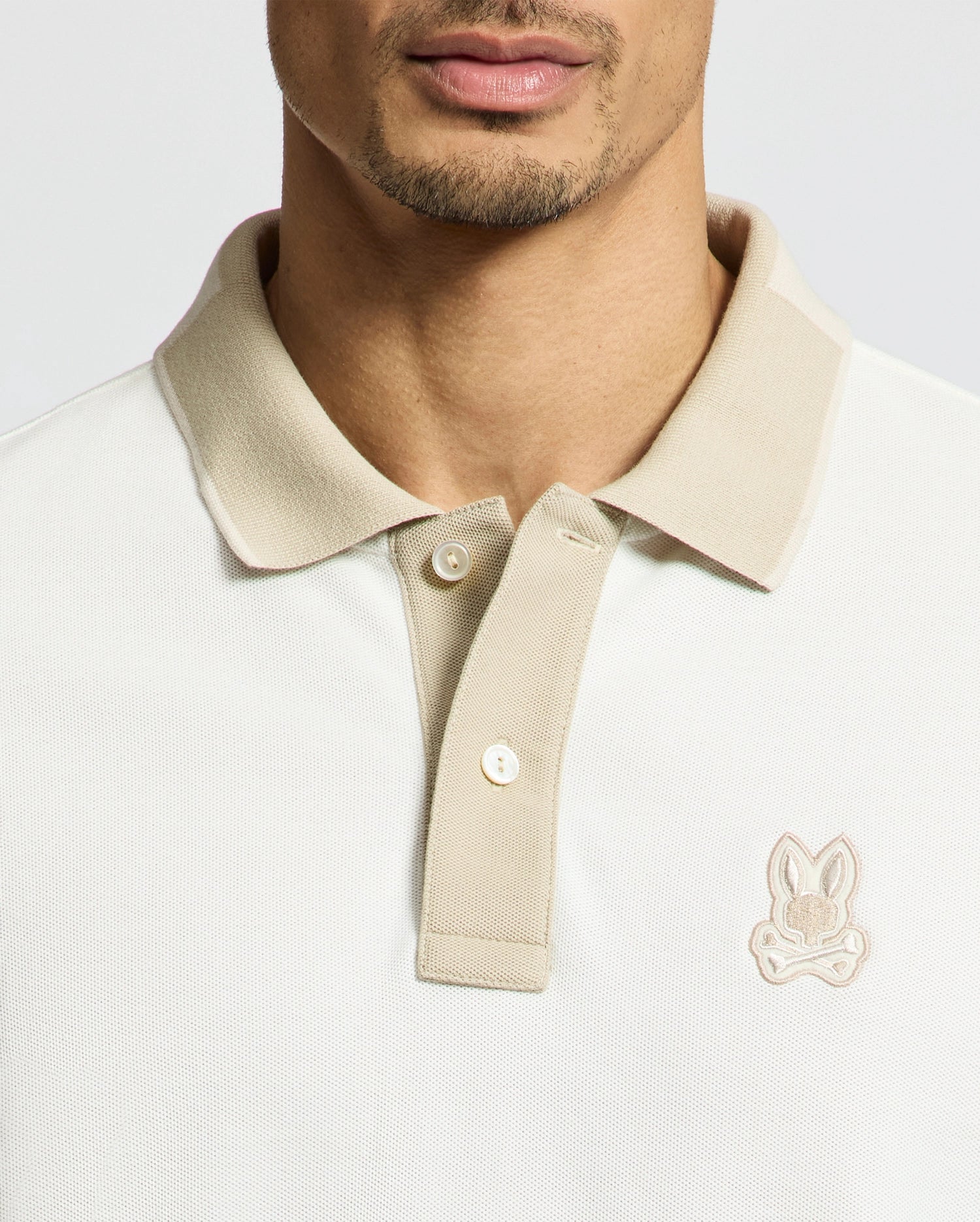 Close-up of a man wearing the Psycho Bunny MENS NEW CASTLE PIQUE POLO (B6K487F200), a slim fit light beige Pima Cotton polo with contrast collar detailing and a rabbit logo on the left chest, showing his neck, chin, and jawline.