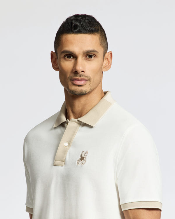 A man with short dark hair and trimmed facial hair poses against a plain light background wearing the Psycho Bunny MENS NEW CASTLE PIQUE POLO (B6K487F200): a slim fit white polo with beige collar, contrast details, and embroidered bunny logo on the chest.