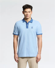A person poses in a Psycho Bunny MENS NEW CASTLE PIQUE POLO - B6K487F200, showcasing its slim fit, light blue Pima cotton fabric with an embroidered bunny logo on the chest and dark blue collar/sleeve trim, paired elegantly with beige pants.