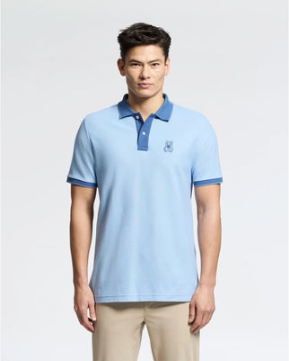 A person poses in a Psycho Bunny MENS NEW CASTLE PIQUE POLO - B6K487F200, showcasing its slim fit, light blue Pima cotton fabric with an embroidered bunny logo on the chest and dark blue collar/sleeve trim, paired elegantly with beige pants.
