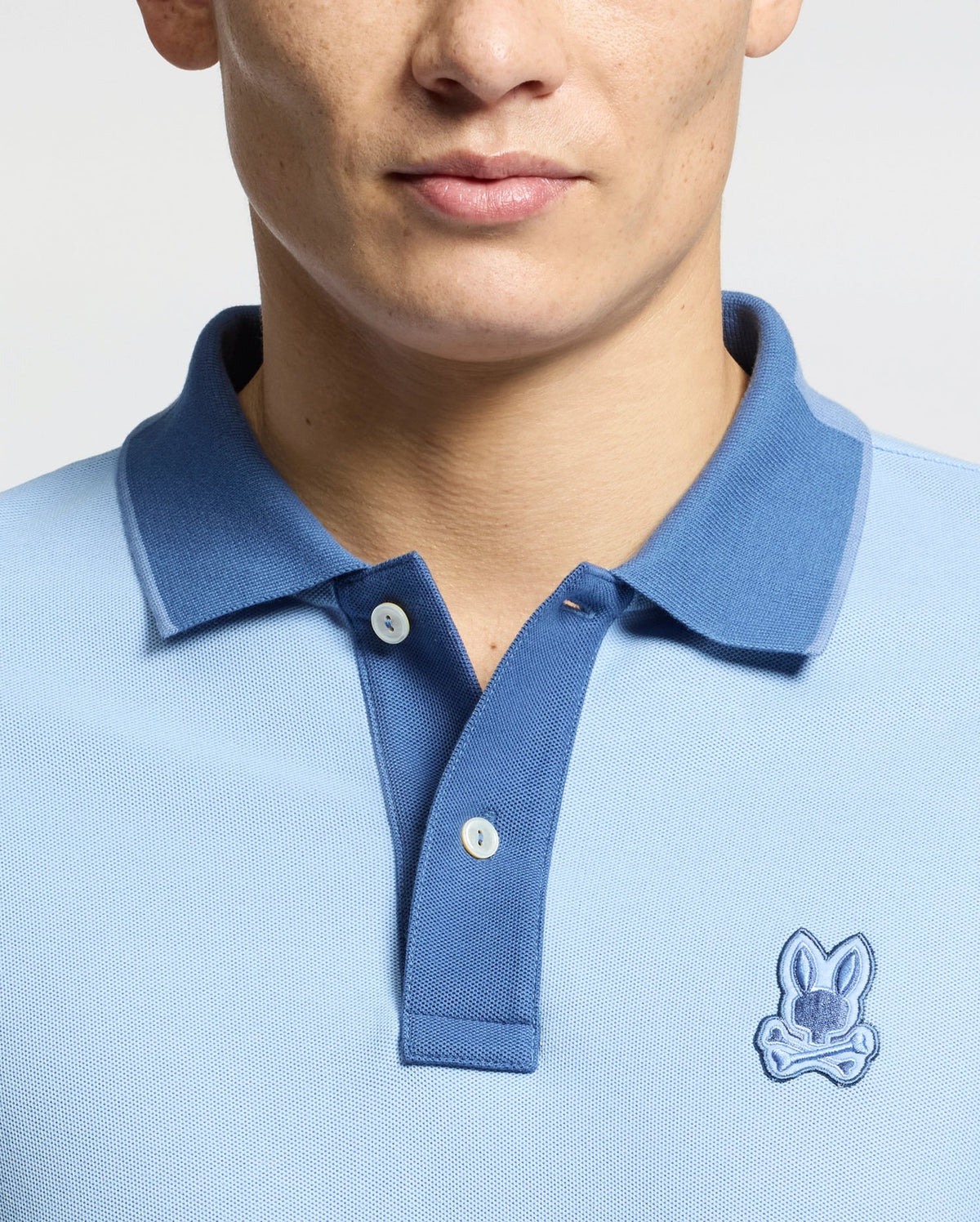 A person is wearing the Psycho Bunny MENS NEW CASTLE PIQUE POLO in light blue with a darker blue collar, featuring an embroidered bunny logo on the chest. Made from Pima cotton, the shirt has two visible buttons. The photo shows only the shirt's upper part and part of the person's face.