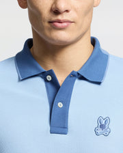 A person is wearing the Psycho Bunny MENS NEW CASTLE PIQUE POLO in light blue with a darker blue collar, featuring an embroidered bunny logo on the chest. Made from Pima cotton, the shirt has two visible buttons. The photo shows only the shirt's upper part and part of the person's face.