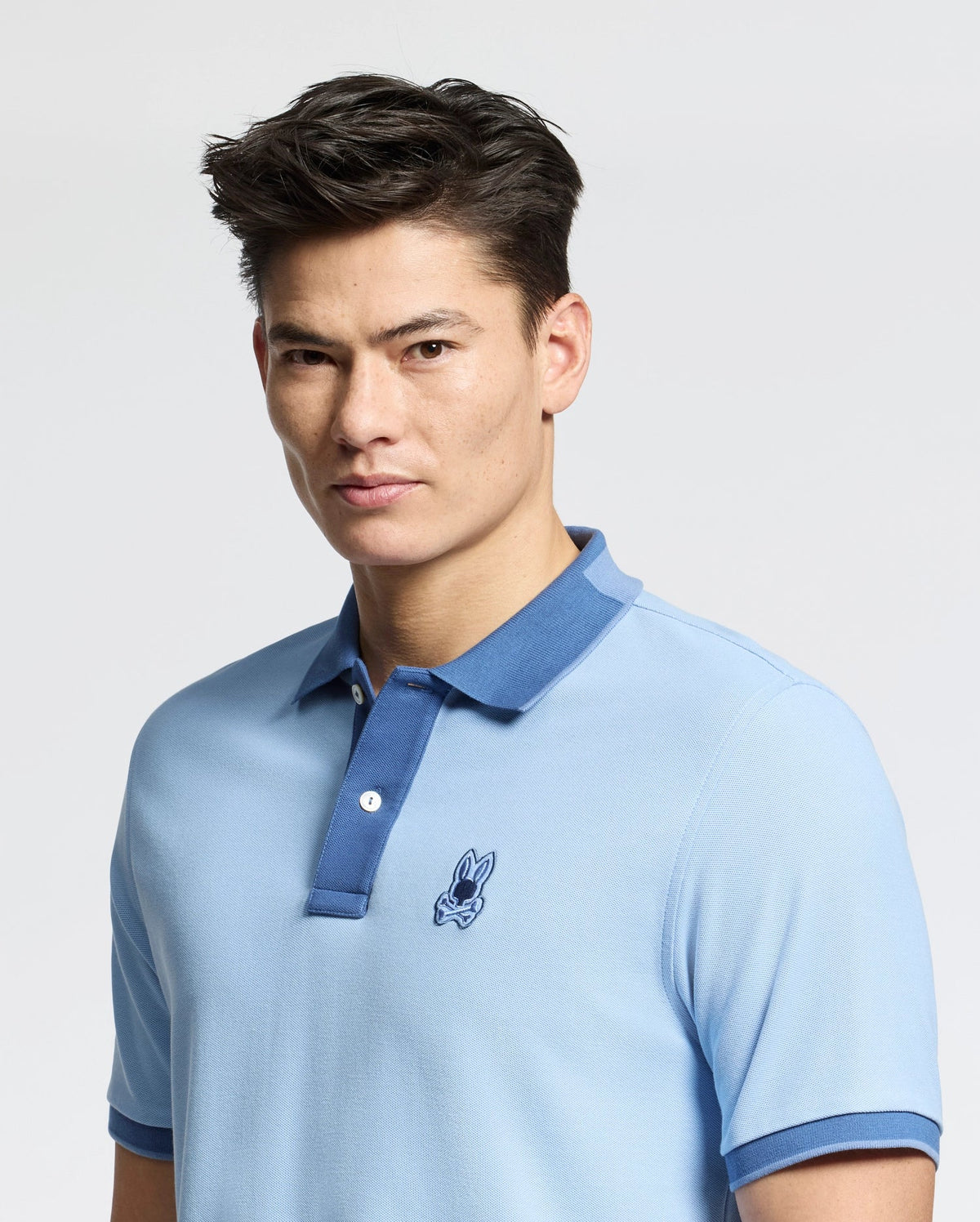 A person with short, dark hair wears a slim fit, light blue MENS NEW CASTLE PIQUE POLO - B6K487F200 from Psycho Bunny. Made from soft Pima cotton, it features a blue collar and an embroidered bunny patch on the chest. They stand against a plain white background, gazing into the camera.