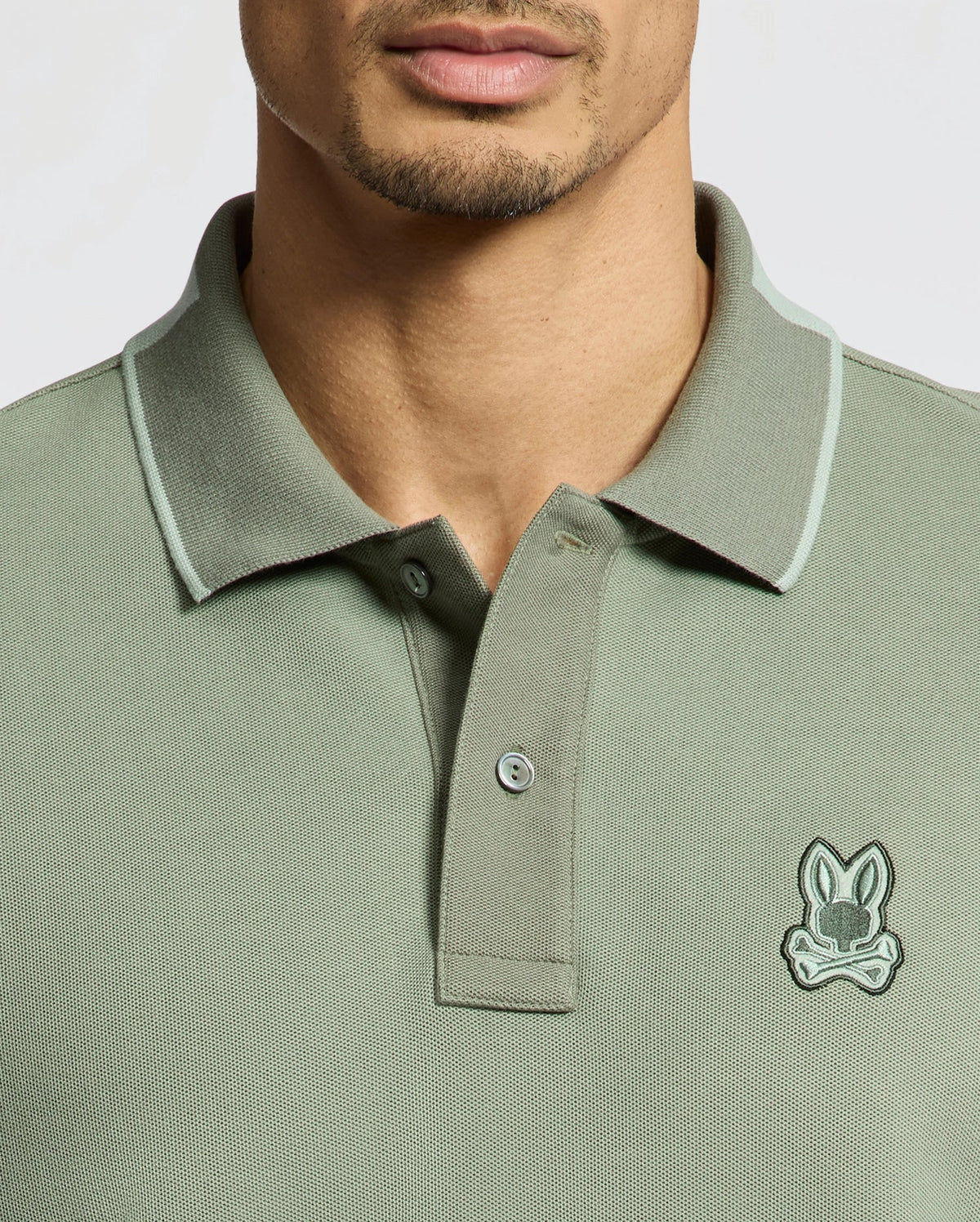 Someone is wearing a Psycho Bunny MENS NEW CASTLE PIQUE POLO - B6K487F200 in green, made of premium Pima Cotton. It features a small embroidered stylized animal logo on the chest, with a collar and button placket on a plain, light background.