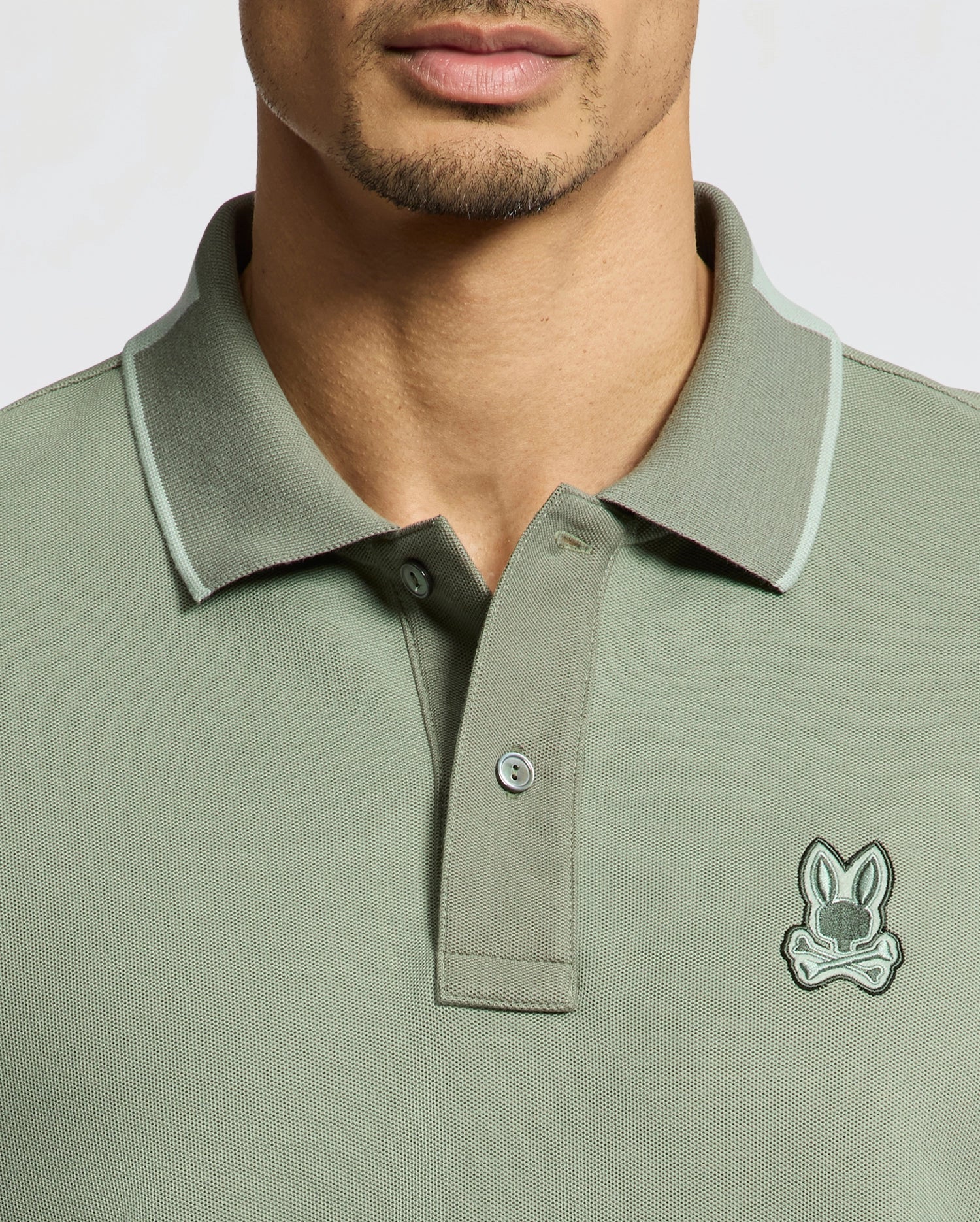 Someone is wearing a Psycho Bunny MENS NEW CASTLE PIQUE POLO - B6K487F200 in green, made of premium Pima Cotton. It features a small embroidered stylized animal logo on the chest, with a collar and button placket on a plain, light background.