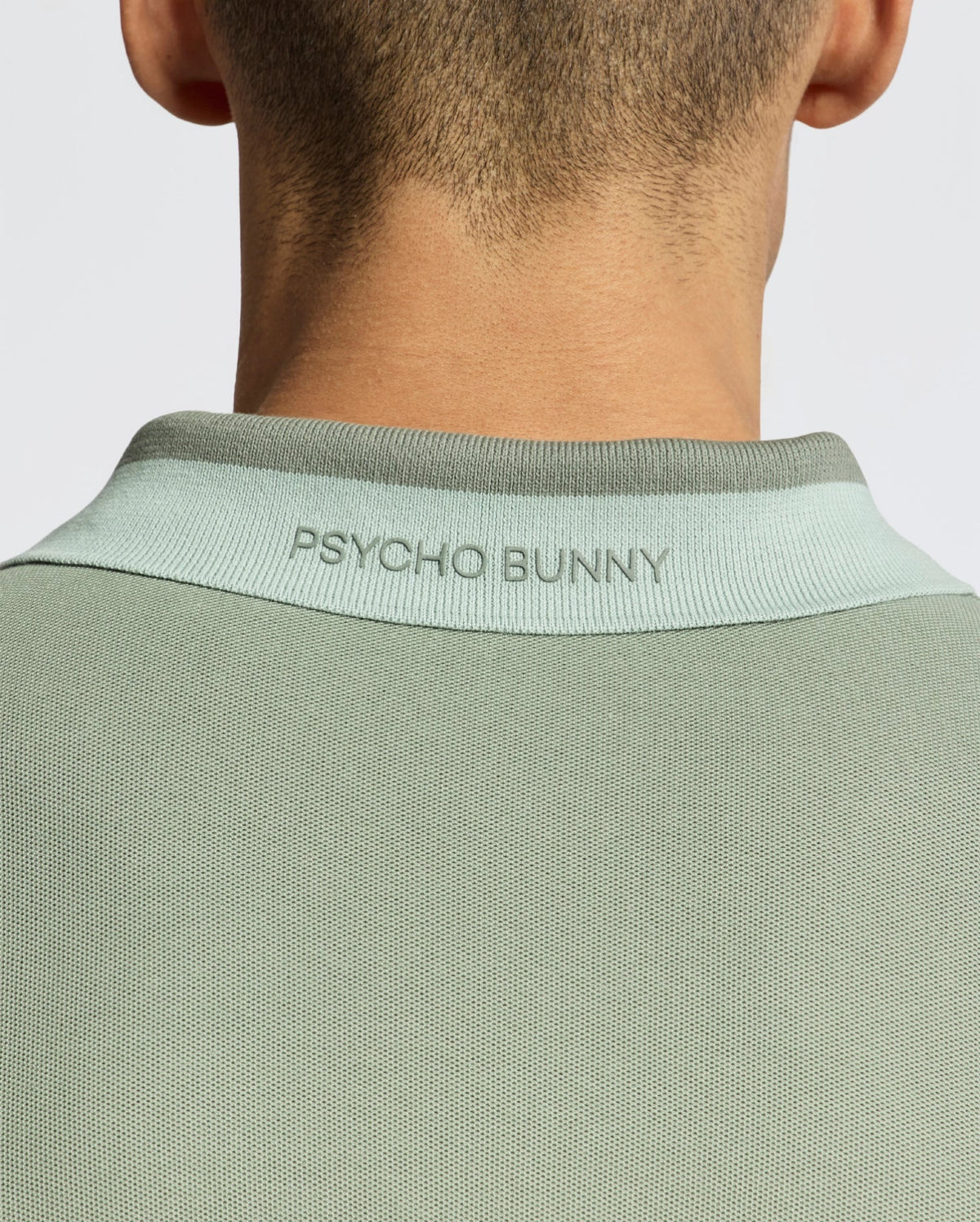Back view of a person wearing the MENS NEW CASTLE PIQUE POLO - B6K487F200 in light green, featuring "PSYCHO BUNNY" on the collar, crafted from soft Pima Cotton with luxury finishes.