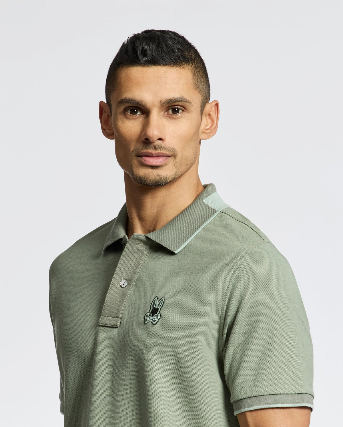 A person stands against a plain white background wearing a soft Pima Cotton Men's New Castle Pique Polo (B6K487F200) by Psycho Bunny in green, featuring a small embroidered logo on the chest.
