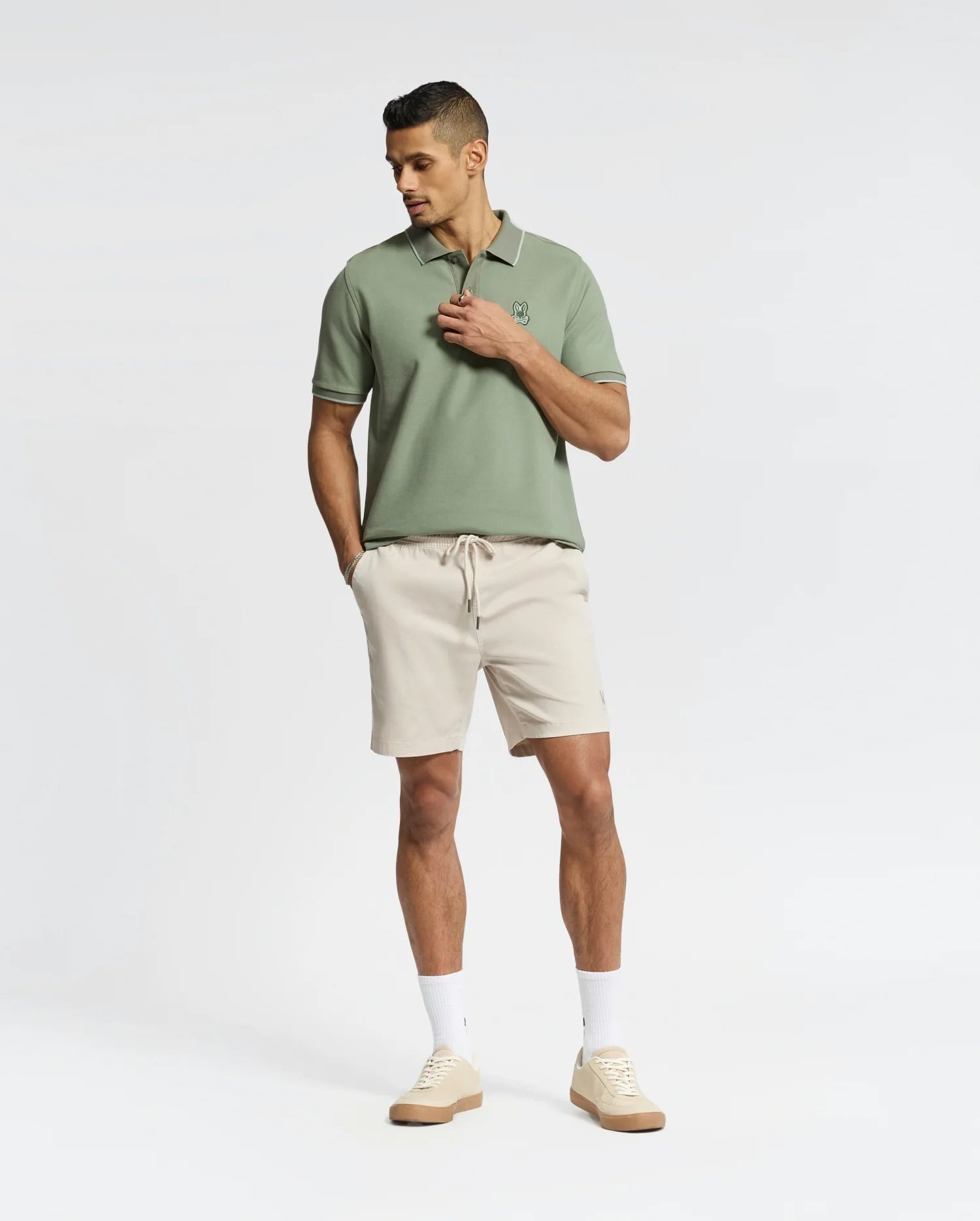 A person stands confidently in a light green Psycho Bunny MENS NEW CASTLE PIQUE POLO - B6K487F200 and beige shorts, hands in pockets, paired with white socks and luxury-finished beige sneakers against a plain white background.