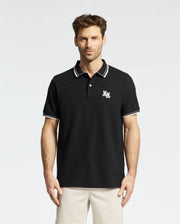 A man stands against a white background wearing the Psycho Bunny MEN'S ELLIOT PIQUE POLO (B6K505F200) in black, featuring an embroidered chest design, white-striped collar and cuffs. Made from Pima cotton, it offers a flawless fit.