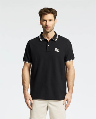 A man stands against a white background wearing the Psycho Bunny MEN'S ELLIOT PIQUE POLO (B6K505F200) in black, featuring an embroidered chest design, white-striped collar and cuffs. Made from Pima cotton, it offers a flawless fit.