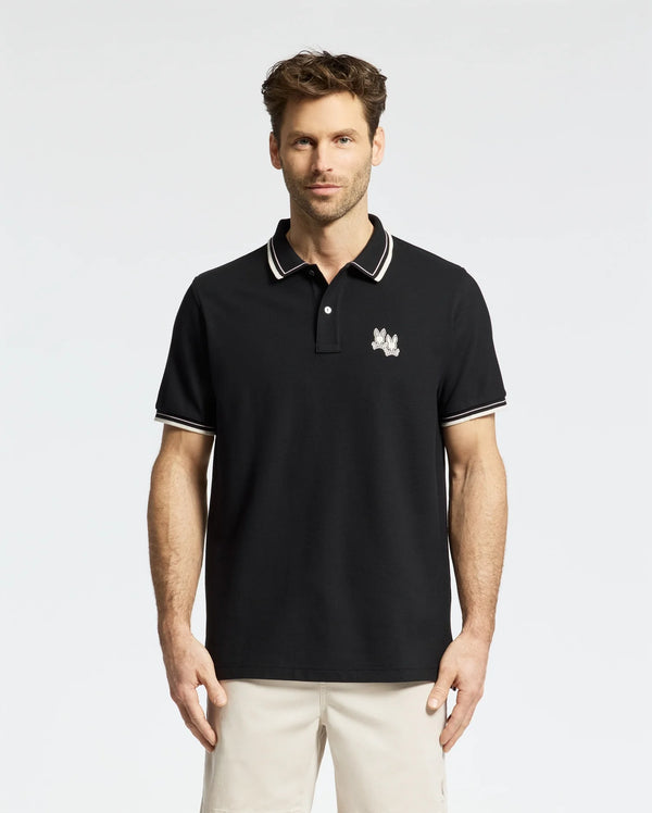 A man stands against a white background wearing the Psycho Bunny MEN'S ELLIOT PIQUE POLO (B6K505F200) in black, featuring an embroidered chest design, white-striped collar and cuffs. Made from Pima cotton, it offers a flawless fit.