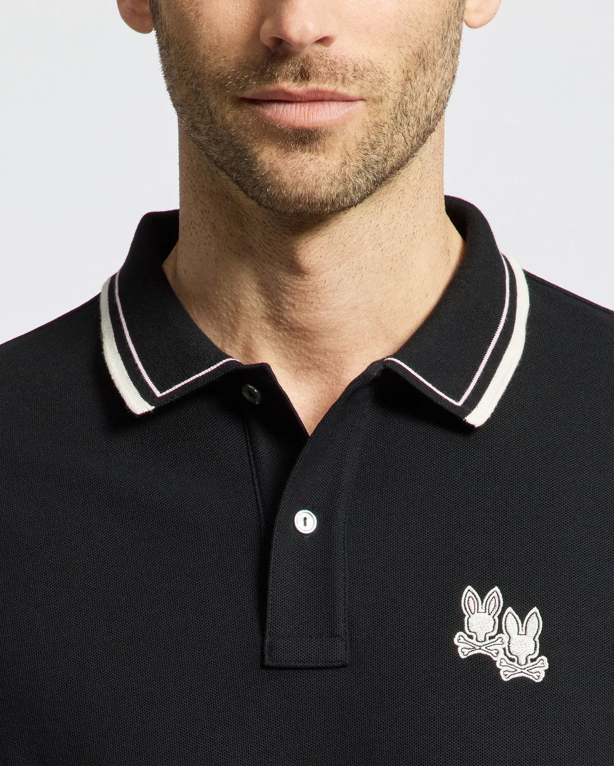 A man sports a Psycho Bunny MENS ELLIOT PIQUE POLO - B6K505F200 in black with a striking white and black striped collar. Crafted from Pima cotton for a perfect fit, it features an embroidered logo of two cartoon rabbits on the chest, set against a plain white background.