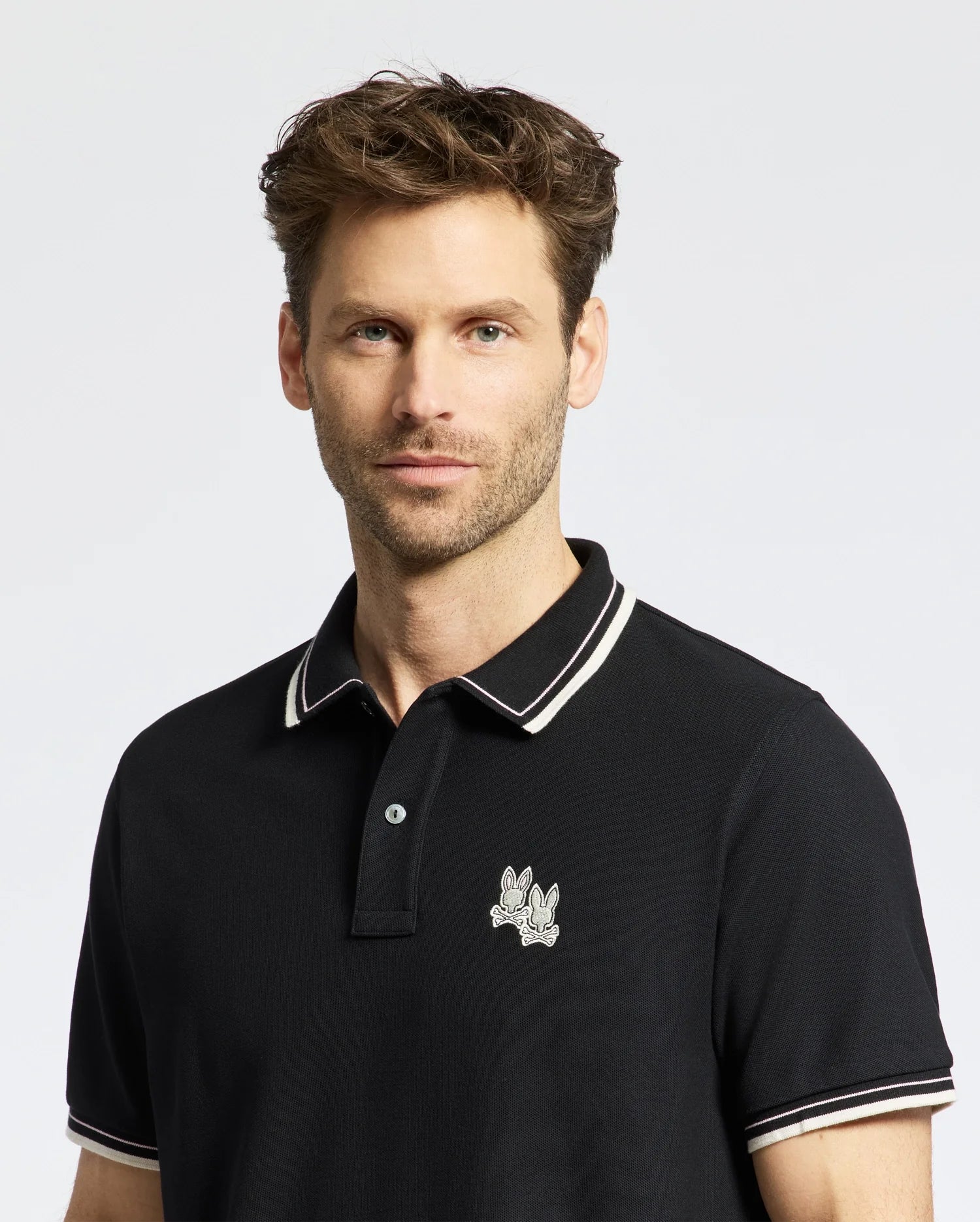 A man with short, wavy hair, a beard, and mustache wears a Psycho Bunny MENS ELLIOT PIQUE POLO - B6K505F200. The black Pima cotton polo features white collar trim and a cartoonish rabbit logo on the chest against a plain light background.