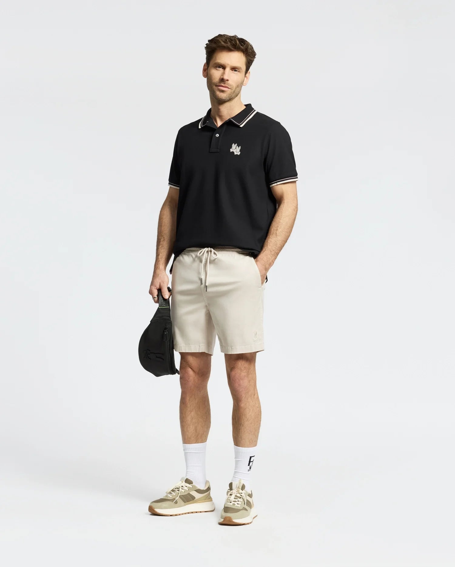 A man exudes confidence in his black MENS ELLIOT PIQUE POLO by Psycho Bunny, featuring bold design elements and a subtle logo. Paired with beige shorts, white socks, and sneakers, his stylish appearance is enhanced by neatly styled hair and a light beard against the plain white backdrop.