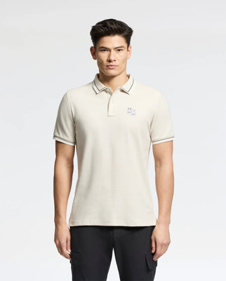 A person is wearing the Psycho Bunny's Men’s Elliot Pique Polo in light beige, featuring short sleeves and an embroidered logo on the chest, set against a white background. They pair it with black pants.